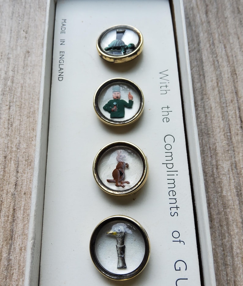 Peut inclure: Ensemble de quatre boutons fantaisie vintage avec des bordures dor&eacute;es. Chaque bouton pr&eacute;sente un design miniature diff&eacute;rent : une tortue, un homme en vert, un lapin et un canard. La bo&icirc;te indique "Made in England" et "With the Compliments of GU".