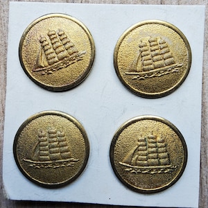 Puede incluir: Cuatro botones de metal de color dorado con un diseño en relieve de un barco con velas. Los botones están dispuestos en un patrón cuadrado sobre un fondo blanco.