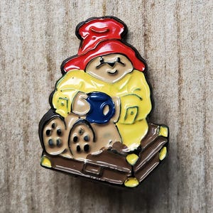 Könnte beinhalten: Eine Emaille-Anstecknadel mit einem Comic-Bären, der einen roten Hut und einen gelben Regenmantel trägt und eine blaue Tasse hält. Der Bär sitzt auf einer braunen Holzplattform.