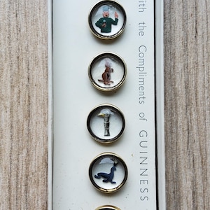 Peut inclure: Un ensemble de six boutons vintage sur le th&egrave;me Guinness, pr&eacute;sent&eacute;s dans une bo&icirc;te blanche. Chaque bouton, avec un bord dor&eacute;, pr&eacute;sente une image miniature diff&eacute;rente : une tortue, un homme, un kangourou, un canard, un phoque et un oiseau. La bo&icirc;te indique "With the Compliments of GUINNESS" et "MADE IN ENGLAND."