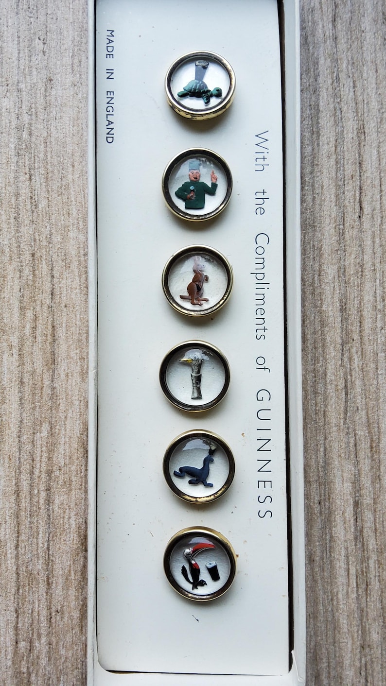 Peut inclure: Un ensemble de six boutons vintage sur le th&egrave;me Guinness, pr&eacute;sent&eacute;s dans une bo&icirc;te blanche. Chaque bouton, avec un bord dor&eacute;, pr&eacute;sente une image miniature diff&eacute;rente : une tortue, un homme, un kangourou, un canard, un phoque et un oiseau. La bo&icirc;te indique "With the Compliments of GUINNESS" et "MADE IN ENGLAND."