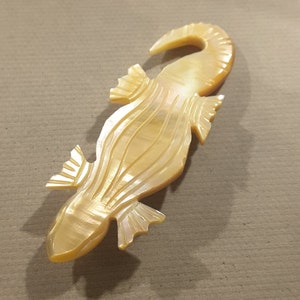 May include: A beige, iridescent, mother-of-pearl lizard brooch. The lizard is carved in a detailed, stylized design with a long tail and a textured body.