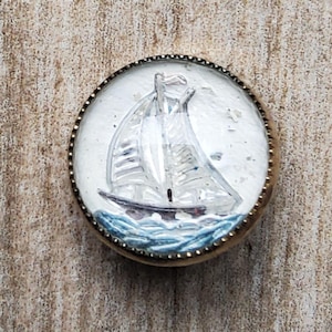 May include: A vintage glass button with a gold rim featuring a white sailboat with blue water. The button is a decorative piece with a nautical theme.