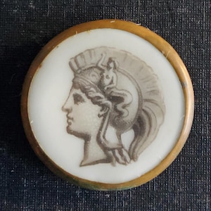 May include: A round, white button with a gold rim. The button features a black and white image of a woman's profile wearing a helmet with a plume.