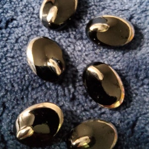 Puede incluir: Seis botones ovalados negros con un borde plateado. Los botones están dispuestos en semicírculo sobre una superficie azul y peluda.