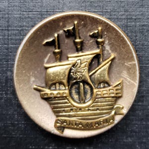 Può includere: Un bottone rotondo in stile antico con un'immagine in rilievo di una nave a vela. La nave è color oro con sartiame e vele dettagliate. La parola "SANTA MARIA" è incisa su uno stendardo. Il bottone ha un bordo color bronzo.