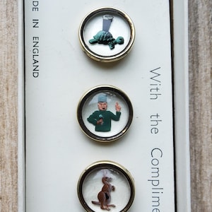 Peut inclure: Ensemble de boutons vintage avec des motifs fantaisistes. Chaque bouton pr&eacute;sente une illustration diff&eacute;rente : une tortue avec un verre, un homme en manteau vert et un kangourou. Les boutons sont sertis dans des cadres en m&eacute;tal dor&eacute;.