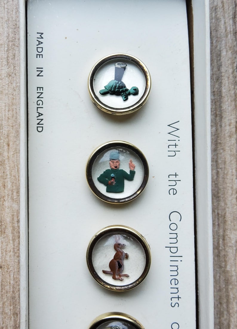 Peut inclure: Ensemble de boutons vintage avec des motifs fantaisistes. Chaque bouton pr&eacute;sente une illustration diff&eacute;rente : une tortue avec un verre, un homme en manteau vert et un kangourou. Les boutons sont sertis dans des cadres en m&eacute;tal dor&eacute;.