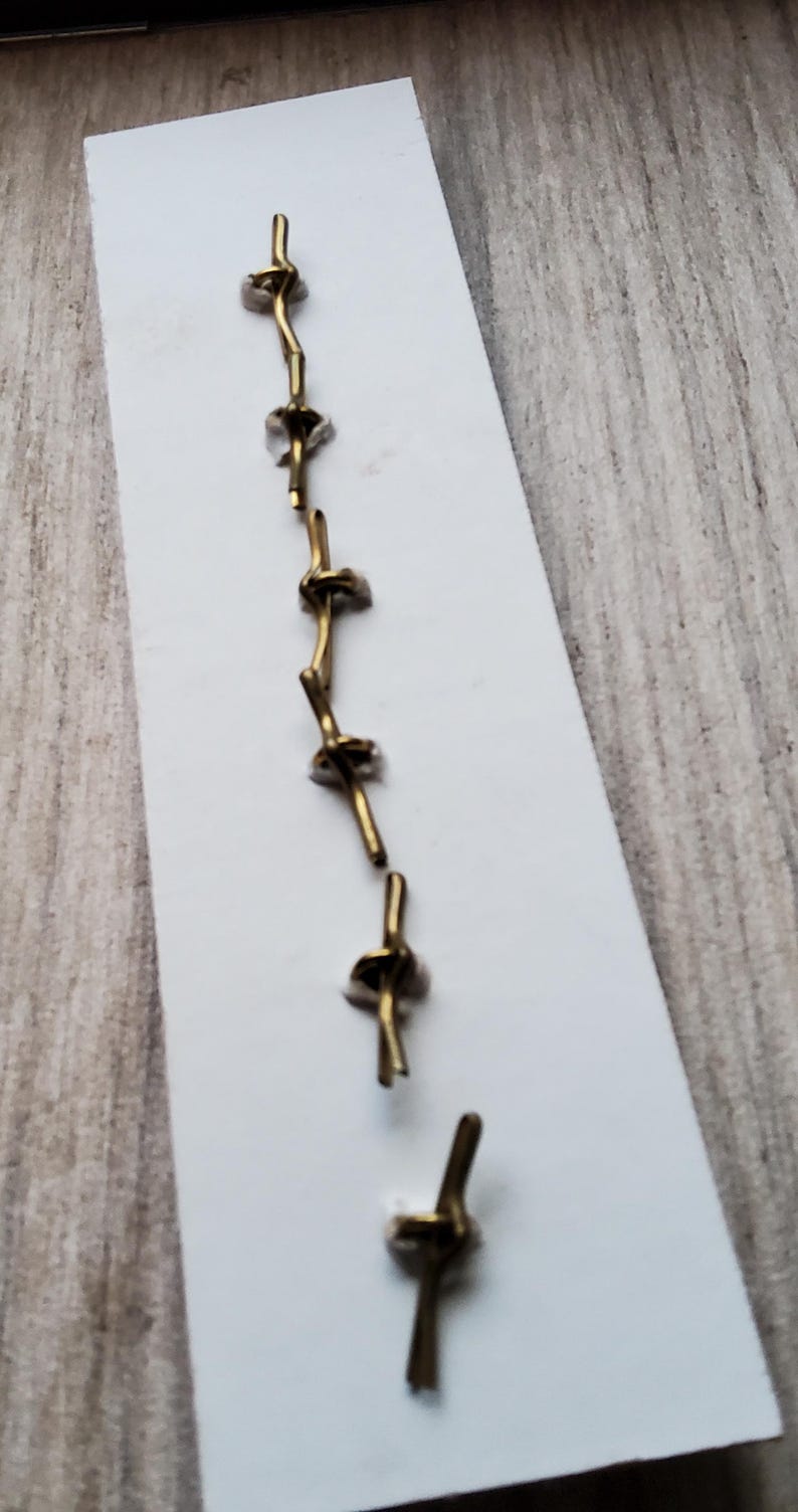 May include: A set of seven gold-colored metal hooks, arranged vertically on a white card. The hooks are designed to be used for hanging items.