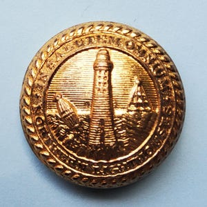 May include: A round, gold-colored button with a detailed nautical design. The central image features a lighthouse, a buoy, and a sailboat, surrounded by text. The button's edge has a rope-like texture.