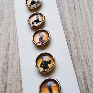 May include: Six round, gold-rimmed buttons with different images inside. The images include a person in a green uniform, a person in a black suit, a person in a brown suit, a person in a black suit, a person in a black suit, and a person in a black suit.