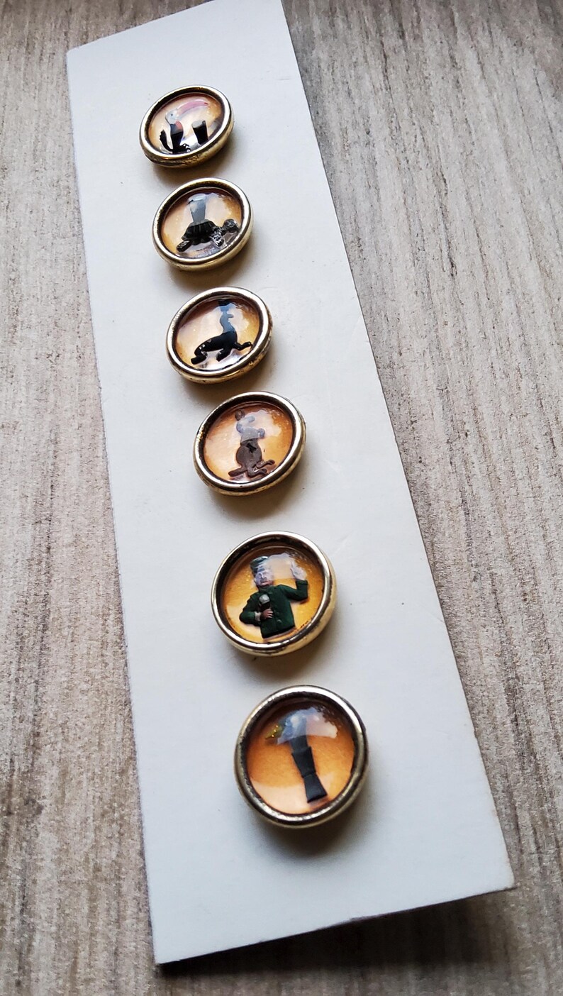 May include: Six round, gold-rimmed buttons with different images inside. The images include a person in a green uniform, a person in a black suit, a person in a brown suit, a person in a black suit, a person in a black suit, and a person in a black suit.