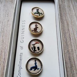 May include: A set of vintage Guinness buttons in a white box. Each button has a gold-colored rim and features a different animal illustration, including a turtle, kangaroo, toucan, and seal. The box reads "the Compliments of Guinness."