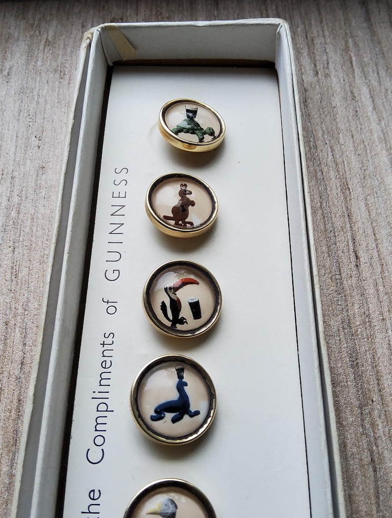 May include: A set of vintage Guinness buttons in a white box. Each button has a gold-colored rim and features a different animal illustration, including a turtle, kangaroo, toucan, and seal. The box reads "the Compliments of Guinness."