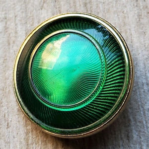 May include: A circular vintage button with a gold-coloured rim and a bright green centre. The centre has a radial pattern, and the button has a dark green outer ring. The button is likely made of metal and glass.