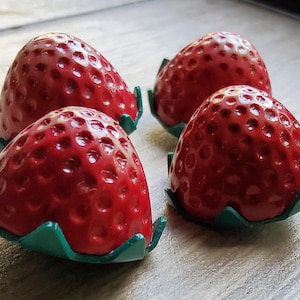 Può includere: Quattro decorazioni a forma di fragola rossa con basi di foglie verdi. Le fragole hanno una finitura lucida e sono disposte su una superficie di colore chiaro. L'immagine è un primo piano.