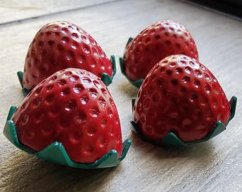 Könnte beinhalten: Vier rote, erdbeerförmige Dekorationen mit grünen Blattbasen. Die Erdbeeren haben eine glänzende Oberfläche und sind auf einer hellen Oberfläche angeordnet. Das Bild ist eine Nahaufnahme.