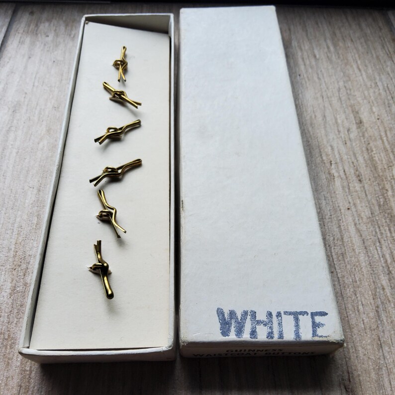 Peut inclure: Un ensemble de six boutons de gilet dor&eacute;s dans une bo&icirc;te blanche. Les boutons sont en forme de n&oelig;ud. La bo&icirc;te est ouverte, r&eacute;v&eacute;lant les boutons dans un int&eacute;rieur blanc. La bo&icirc;te est &eacute;tiquet&eacute;e "WHITE" en texte bleu.