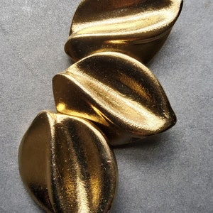 May include: Three gold-colored, leaf-shaped decorative objects arranged vertically. The objects have a smooth, reflective surface, and are positioned against a gray background. The design is abstract and modern.