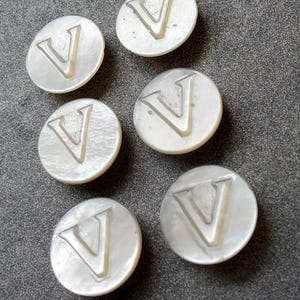 Könnte beinhalten: Sechs weiße Perlmuttknöpfe mit dem Buchstaben "V" auf jedem.