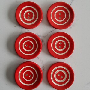 Puede incluir: Un conjunto de seis botones redondos dispuestos en dos columnas. Cada botón es de color rojo brillante con un patrón de anillo circular blanco empotrado y dos pequeños agujeros en el centro para su fijación. Tienen un acabado mate y liso.