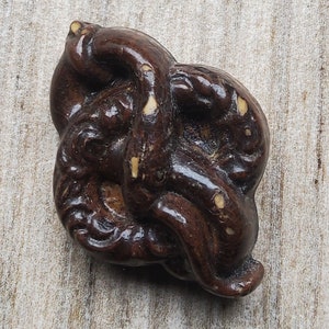 May include: A dark brown, intricately carved wooden object with a swirling, organic design. The object has a smooth, polished surface with lighter-colored accents. The overall shape is rounded and compact, suggesting a decorative or ornamental piece.