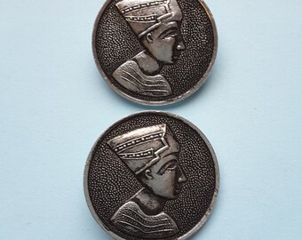 May include: Two silver-toned metal buttons featuring a raised image of a person wearing a crown. The buttons are round and have a textured surface.