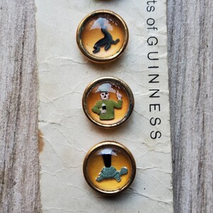 May include: A collection of vintage Guinness-themed buttons. Each button features a different illustration within a gold-toned frame. The illustrations include a seal, a man in a green uniform, and a turtle. The text "ments of GUINNESS" is visible.