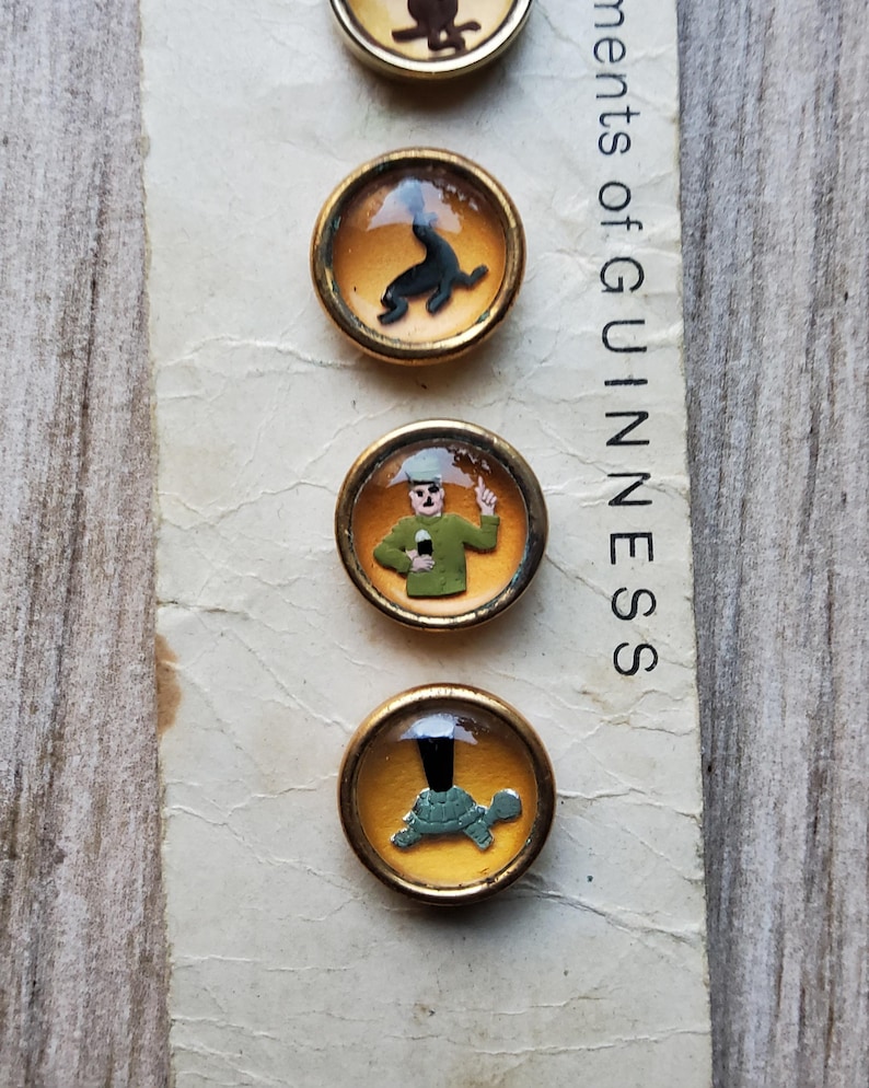 May include: A collection of vintage Guinness-themed buttons. Each button features a different illustration within a gold-toned frame. The illustrations include a seal, a man in a green uniform, and a turtle. The text "ments of GUINNESS" is visible.