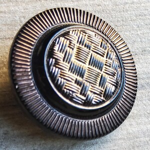 Puede incluir: Un botón negro con un patrón tejido y un anillo exterior plateado.