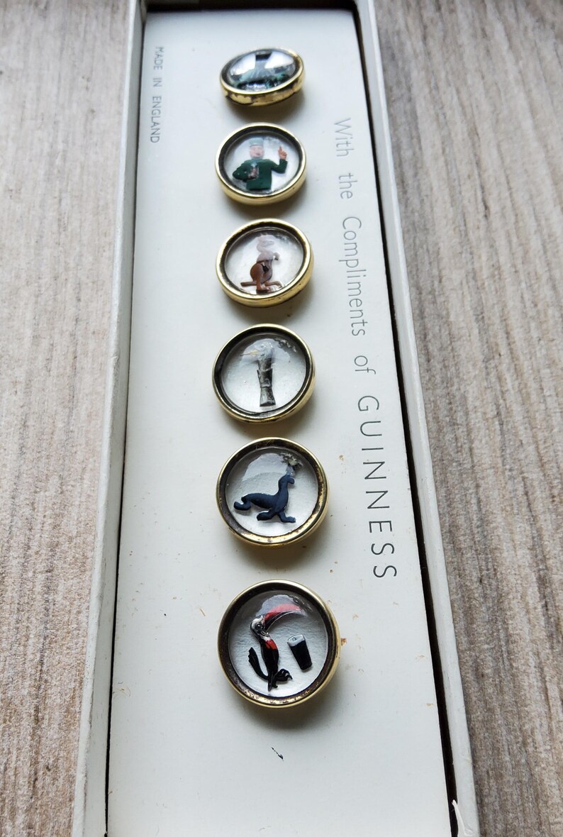 Peut inclure: Un ensemble de six &eacute;pingles promotionnelles Guinness vintage avec des bords dor&eacute;s. Chaque &eacute;pingle pr&eacute;sente une image diff&eacute;rente sous un couvercle transparent en forme de d&ocirc;me. Les &eacute;pingles sont pr&eacute;sent&eacute;es dans une bo&icirc;te blanche avec le texte "With the Compliments of GUINNESS".