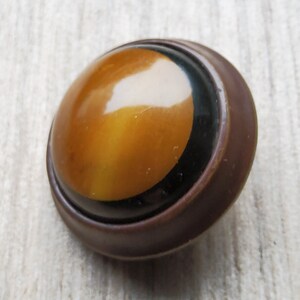 Puede incluir: Un botón redondo de color marrón con un borde negro y un centro amarillo. El botón tiene un acabado brillante.