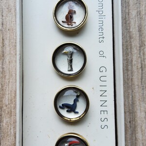 Peut inclure: Ensemble de boutons Guinness vintage avec divers motifs animaliers dans des cadres ronds et transparents de couleur dor&eacute;e. Le texte "Compliments of GUINNESS" est imprim&eacute; verticalement sur le c&ocirc;t&eacute; droit de la bo&icirc;te.