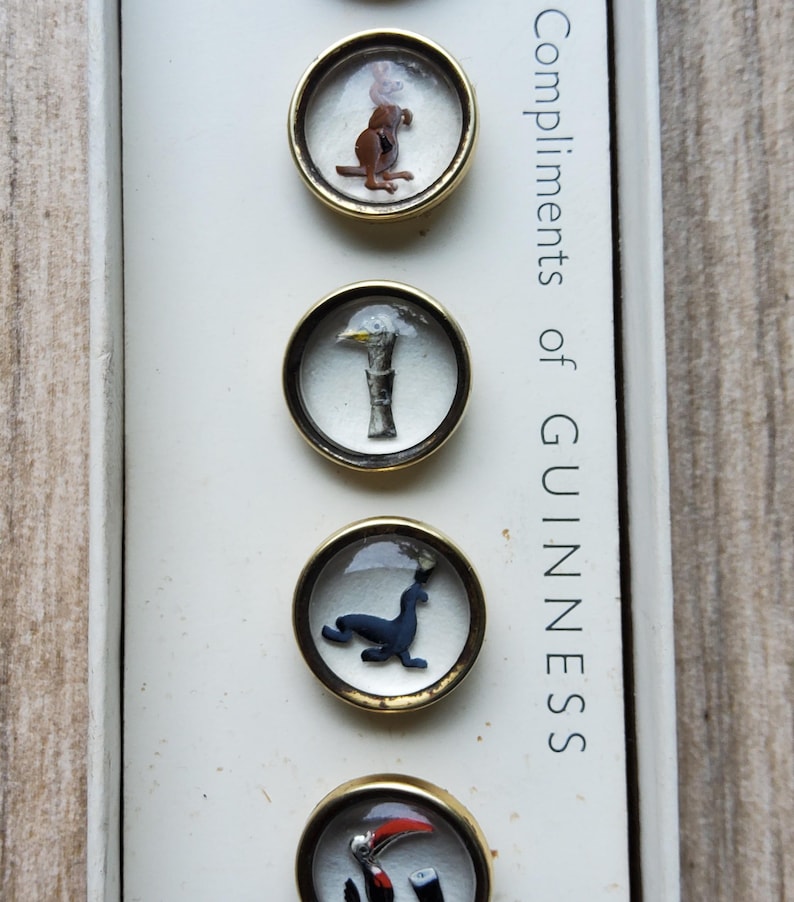 Peut inclure: Ensemble de boutons Guinness vintage avec divers motifs animaliers dans des cadres ronds et transparents de couleur dor&eacute;e. Le texte "Compliments of GUINNESS" est imprim&eacute; verticalement sur le c&ocirc;t&eacute; droit de la bo&icirc;te.