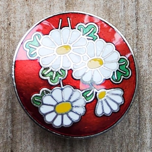 May include: Round, red button with a silver edge, featuring white daisies with yellow centers and green leaves. The floral design is set against a vibrant red background, creating a striking contrast. This decorative button is suitable for various craft projects.