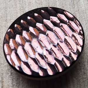 Peut inclure: Bouton ovale avec une surface texturée. Le bouton présente un motif de formes hexagonales superposées dans des tons de marron, de cuivre et de rose. Le bord du bouton est marron foncé.