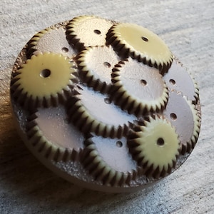 May include: A brown and cream button with a design of overlapping gears. The button has a textured surface and a raised edge.