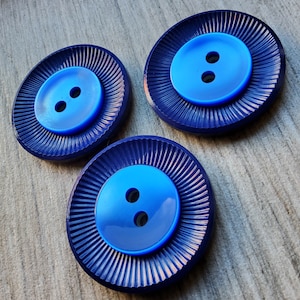 May include: Three blue plastic buttons with two holes each. The buttons have a dark blue outer ring with a lighter blue centre. The buttons are on a wooden surface.