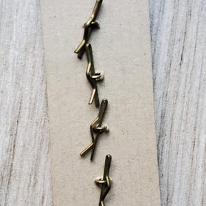 May include: A collection of small, brass-colored metal fasteners arranged on a rectangular piece of cardboard. The fasteners have a unique, bent design, and are likely used for crafts or jewelry making.
