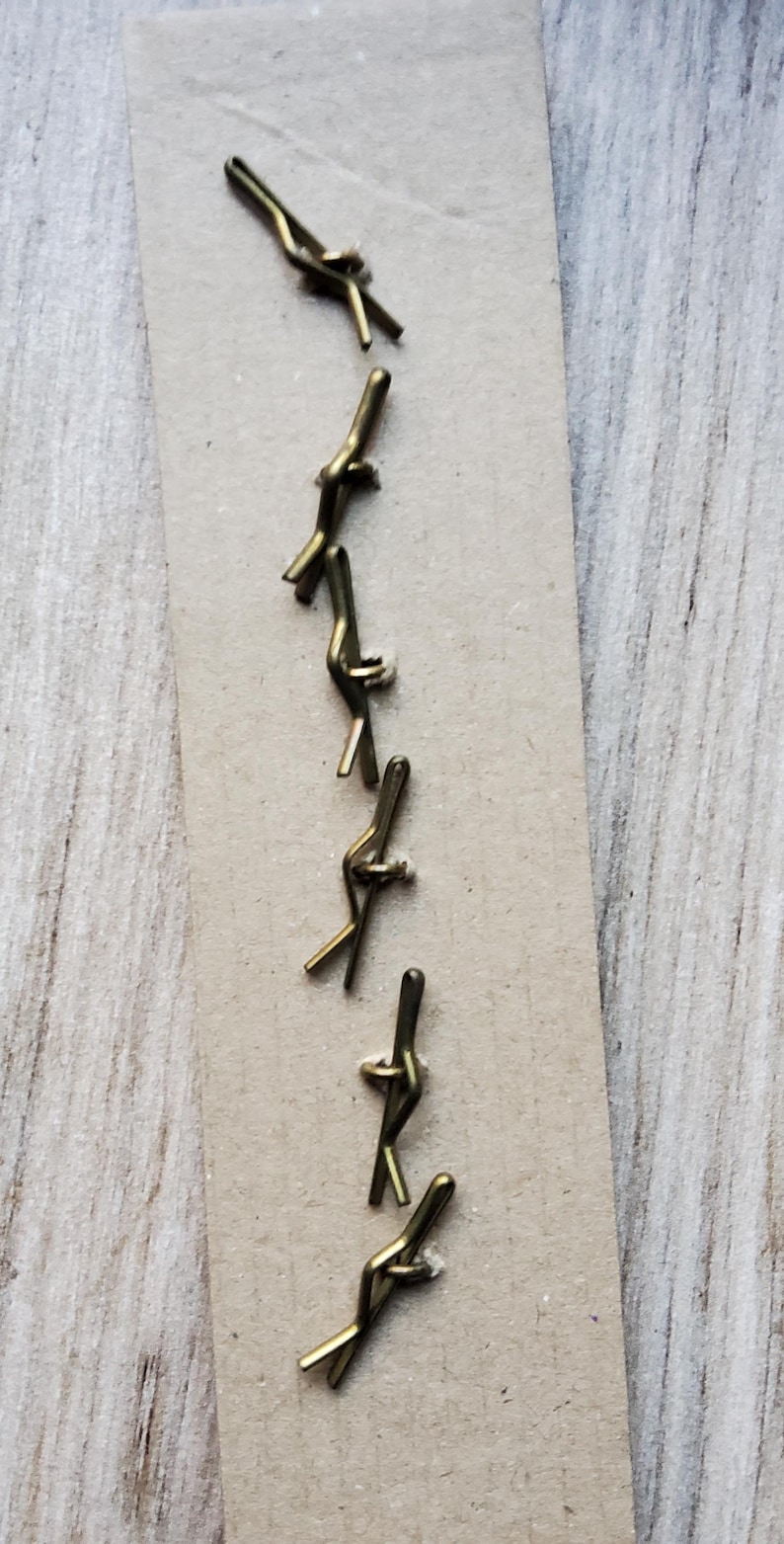 May include: A collection of small, brass-colored metal fasteners arranged on a rectangular piece of cardboard. The fasteners have a unique, bent design, and are likely used for crafts or jewelry making.