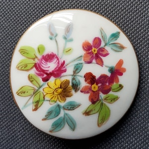 May include: A round, white decorative button with a hand-painted floral design. The flowers are in shades of pink, red, yellow, and orange, with green leaves. The button has a gold-colored rim.