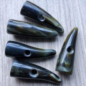 May include: Five horn-shaped toggle buttons with a dark green and brown marbled pattern. Each button has a hole for sewing. The buttons are arranged on a light wood surface.