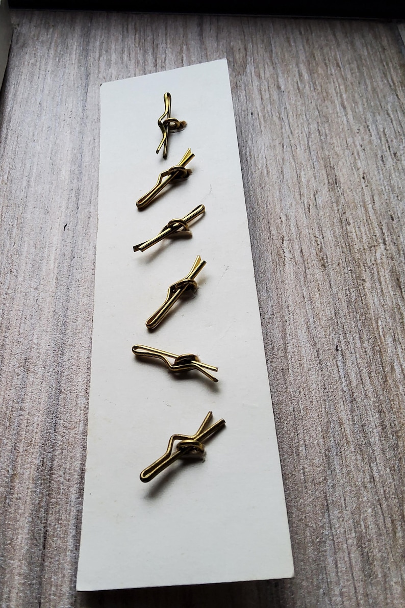 May include: Six vintage gold-toned tie clips arranged on a white card. The tie clips have a simple, elegant design with a knot detail. The background is a light-colored wooden surface, providing a neutral backdrop for the accessories.