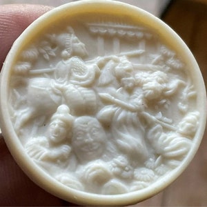 Könnte beinhalten: Ein runder Knopf aus weißem Keramik mit einem erhabenen Design einer Szene mit mehreren Figuren. Die Figuren sind in einem weißen Reliefdesign und scheinen sich in einer Kampfszene zu befinden.
