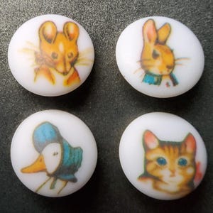 Könnte beinhalten: Vier weiße, runde Knöpfe mit farbenfrohen Illustrationen von Märchenfiguren. Die Bilder zeigen eine Maus, ein Kaninchen, eine Ente und eine Katze. Die Illustrationen sind im Vintage-Stil gehalten, mit sanften Farben und detaillierten Merkmalen.