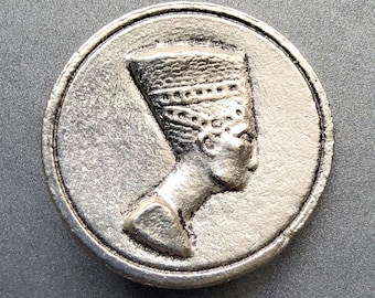 May include: Silver-toned metal coin featuring a raised profile of an Egyptian queen wearing a headdress.