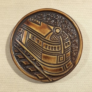 May include: A brown ceramic coaster featuring a detailed relief design of a train engine. The train is brown and has a black and white design.