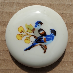 May include: A round, off-white button featuring a colorful illustration of two bluebirds perched on a branch with yellow flowers and green leaves. The birds have black beaks and accents of brown and white.