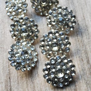 Peut inclure: Gros plan de plusieurs boutons argentés ornés de strass. Chaque bouton présente un strass central plus grand entouré de plus petits, créant un motif floral ou en forme d'éclat d'étoile. Les boutons sont disposés sur une surface claire et texturée.
