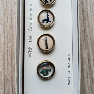 May include: A set of four vintage round buttons with gold-colored rims. Each button features a different whimsical illustration: an eagle, a seal, a duck, and a man. The text "With the Compliments of" and "MADE IN ENGLAND" are also visible.